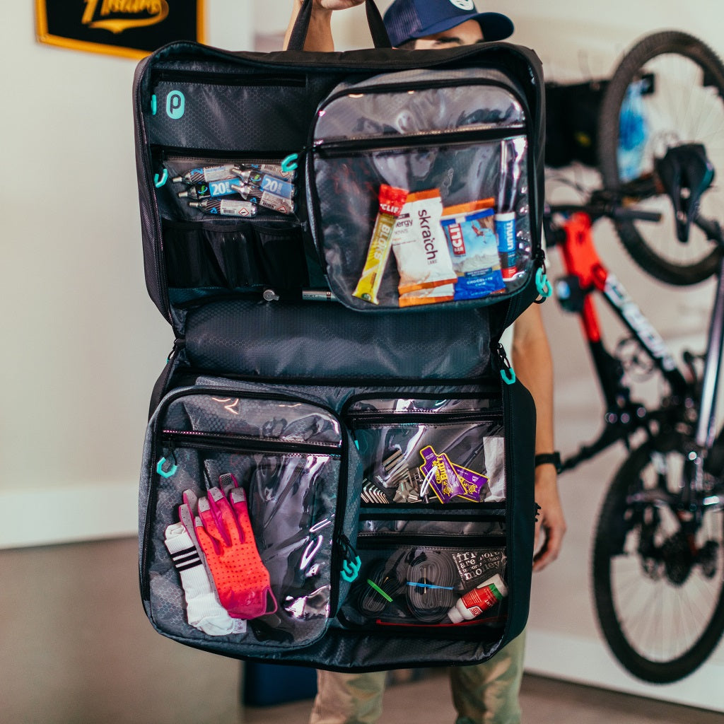 Parc cycling gear bag held up by male in garage with bikes hanging in background. Contents of cycling kit bag visible through clear plastic pockets.