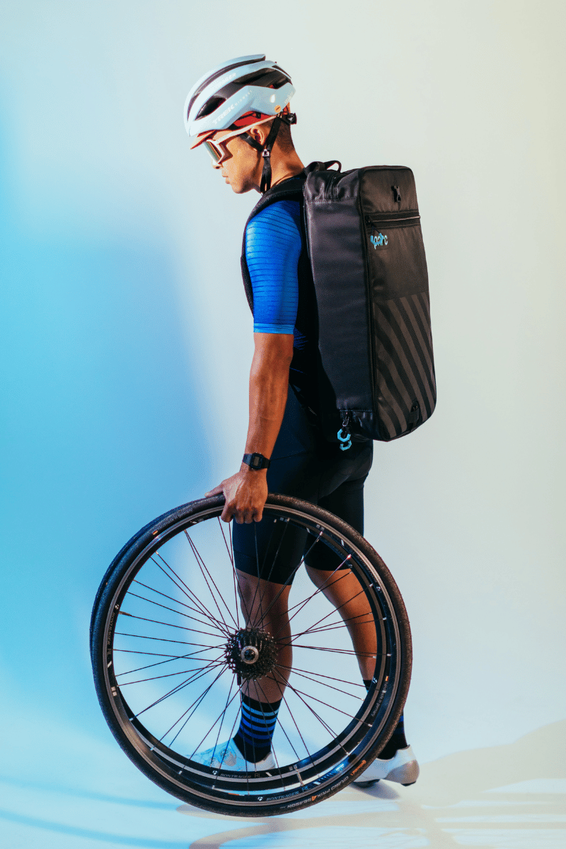 Male cyclist in helmet sunglasses and cycling shoes wears Parc cycling gear bag on his back and holds pair of cycling wheels in left hand.