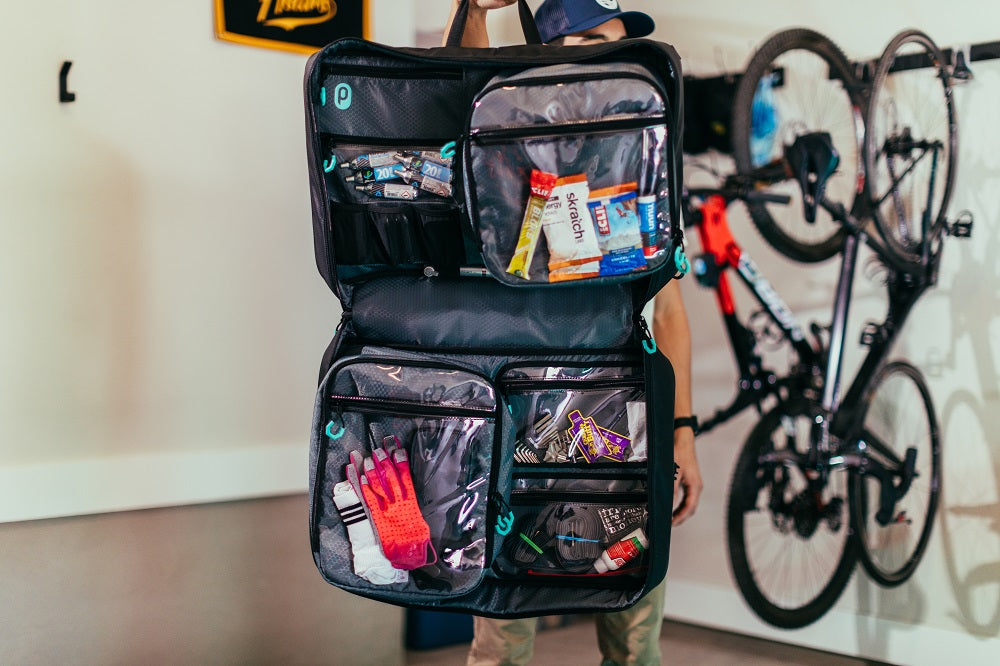 Man standing in garage holding unzipped Parc cycling gear bag in front of his body. Cycling gear and cycling kit visible in Bag. Bikes hang in background.
