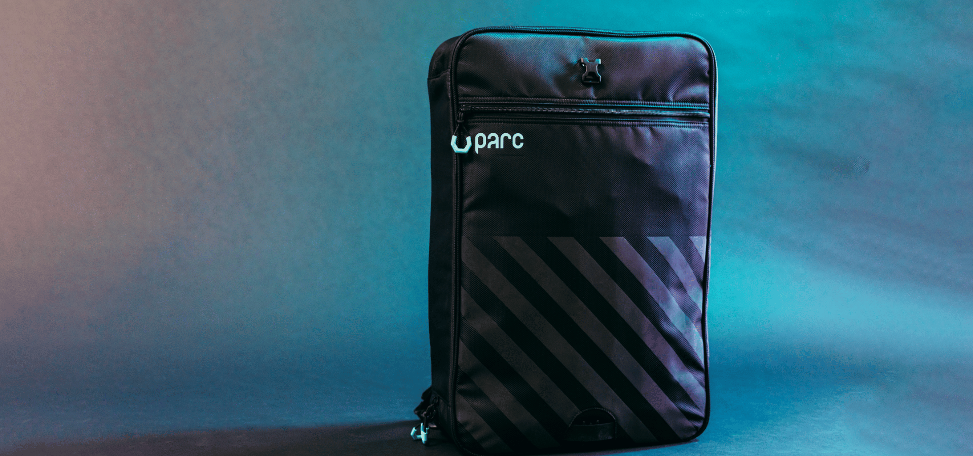 Black Parc cycling gear bag with turquoise logo and zipper pulls against moody blue background. Reflective diagonal stripes on bottom half of exterior front of the cycling kit bag.