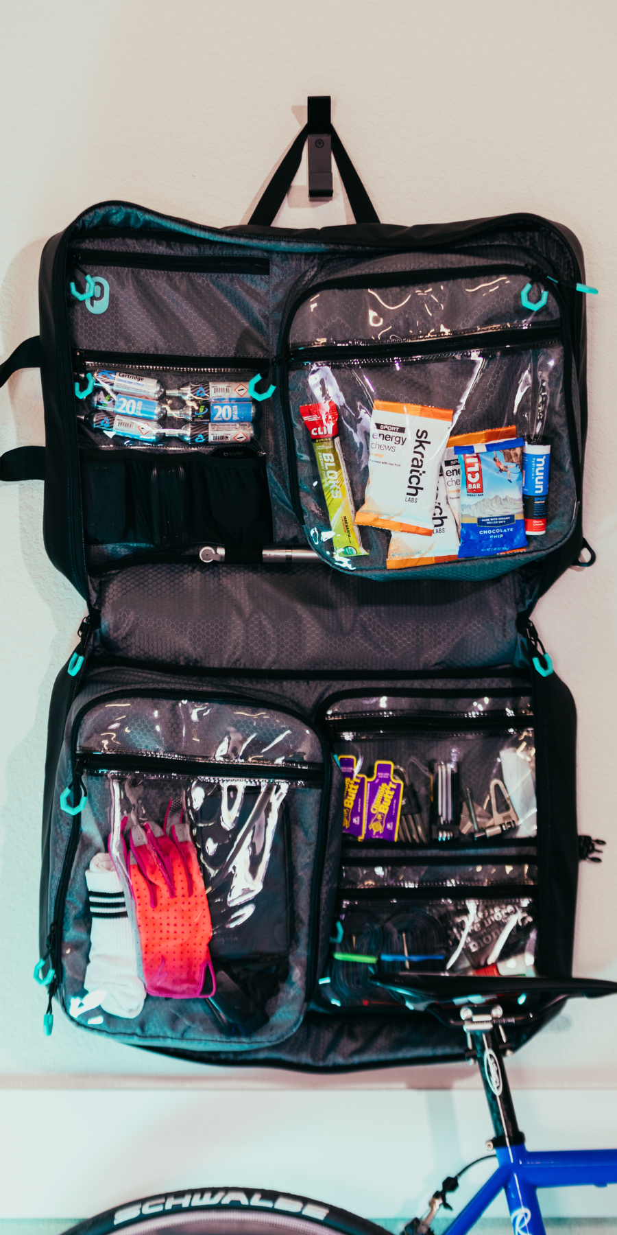 Parc cycling gear bag loaded with cycling kit and cycling gear hangs on wall from hook. Road bike visible leaning against wall in front of cycling bag.
