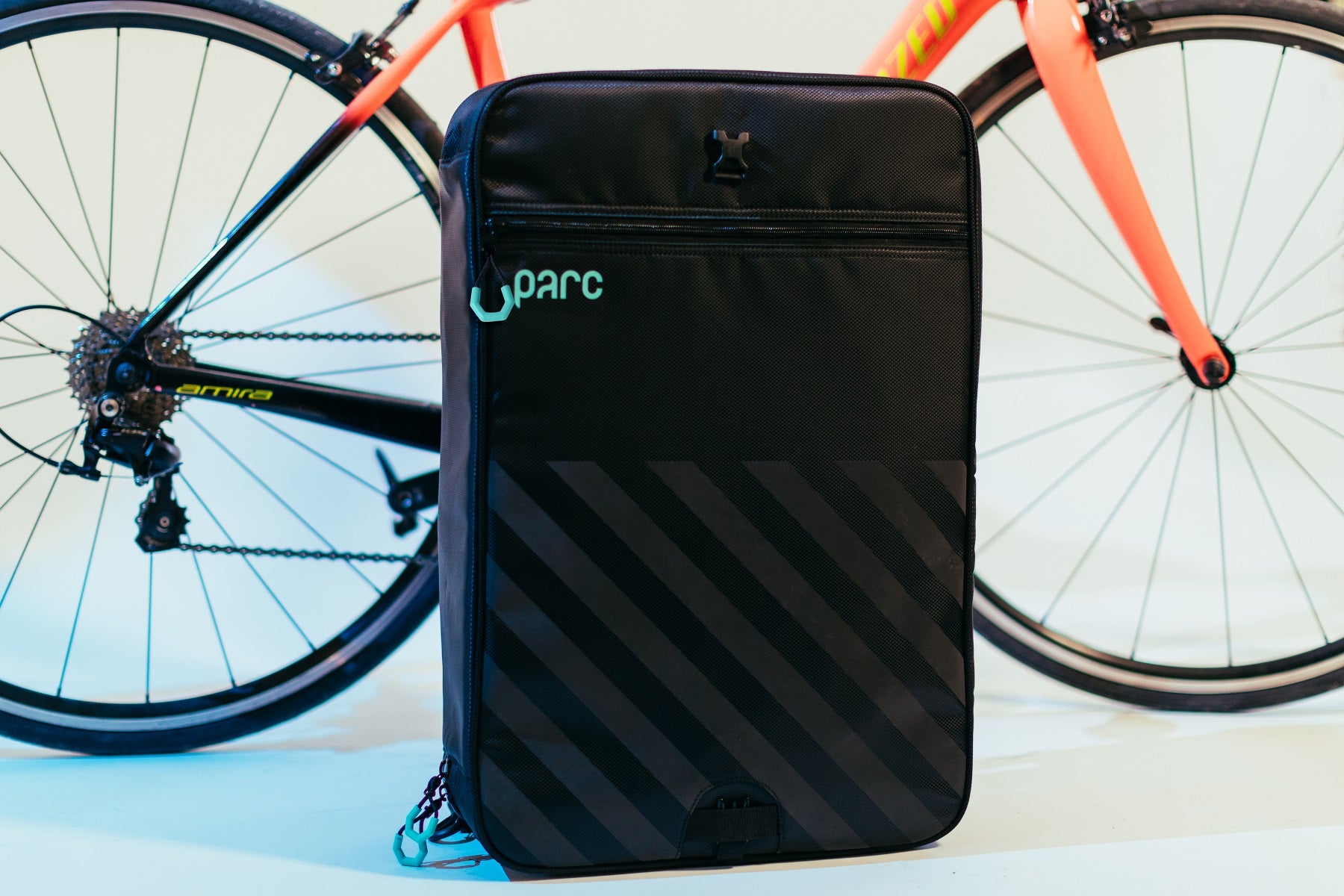 Black Parc cycling kit bag with turquoise logo and zipper pulls is zipped up and standing upright in front of road bike.