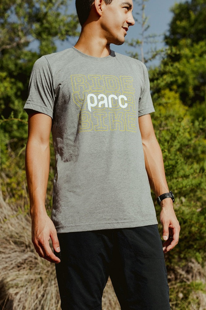 Parc model standing outside wearing grey short sleeve t-shirt. T-shirt illustration on the front chest shows the words ride your bike stacked in a yellow outlined font with a white parc logo over the top.
