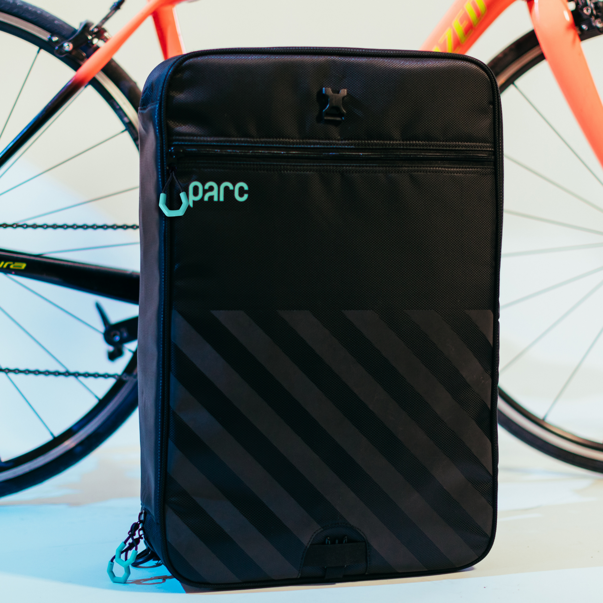 Parc cycling gear bag zipped up and standing upright in front of a road bike. A black bike gear bag with turquoise Parc logo and zipper pulls and black reflective stripes.