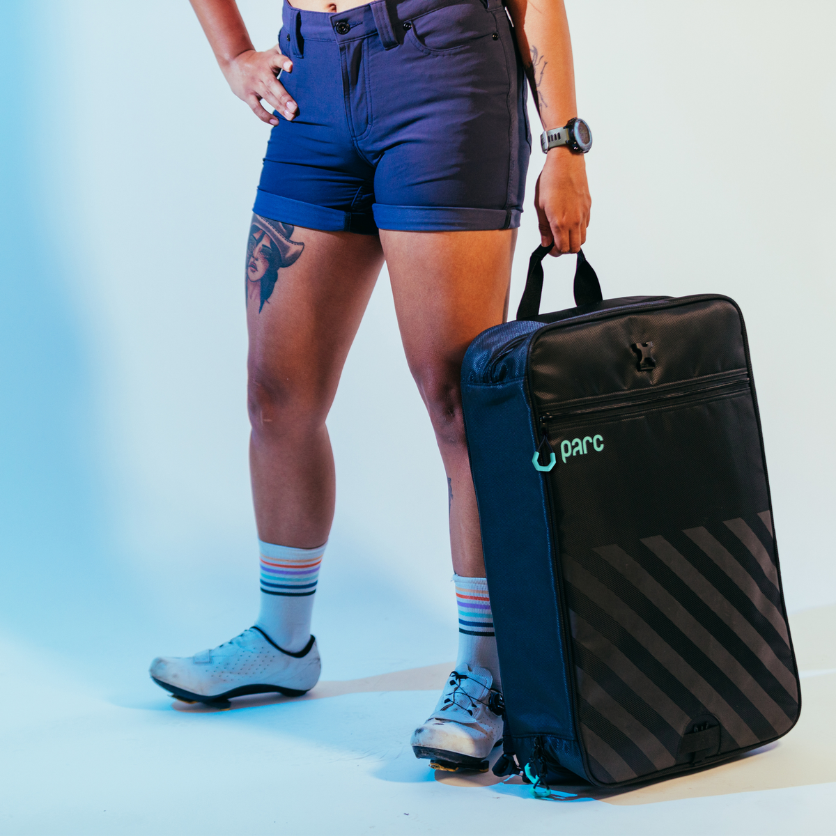 Woman standing in cycling shoes holding Parcs cycling gear bag at her side. Cycling kit bag is zipped up. Black cycling gear bag with turquoise accents and diagonal reflective stripes on bottom half of exterior.