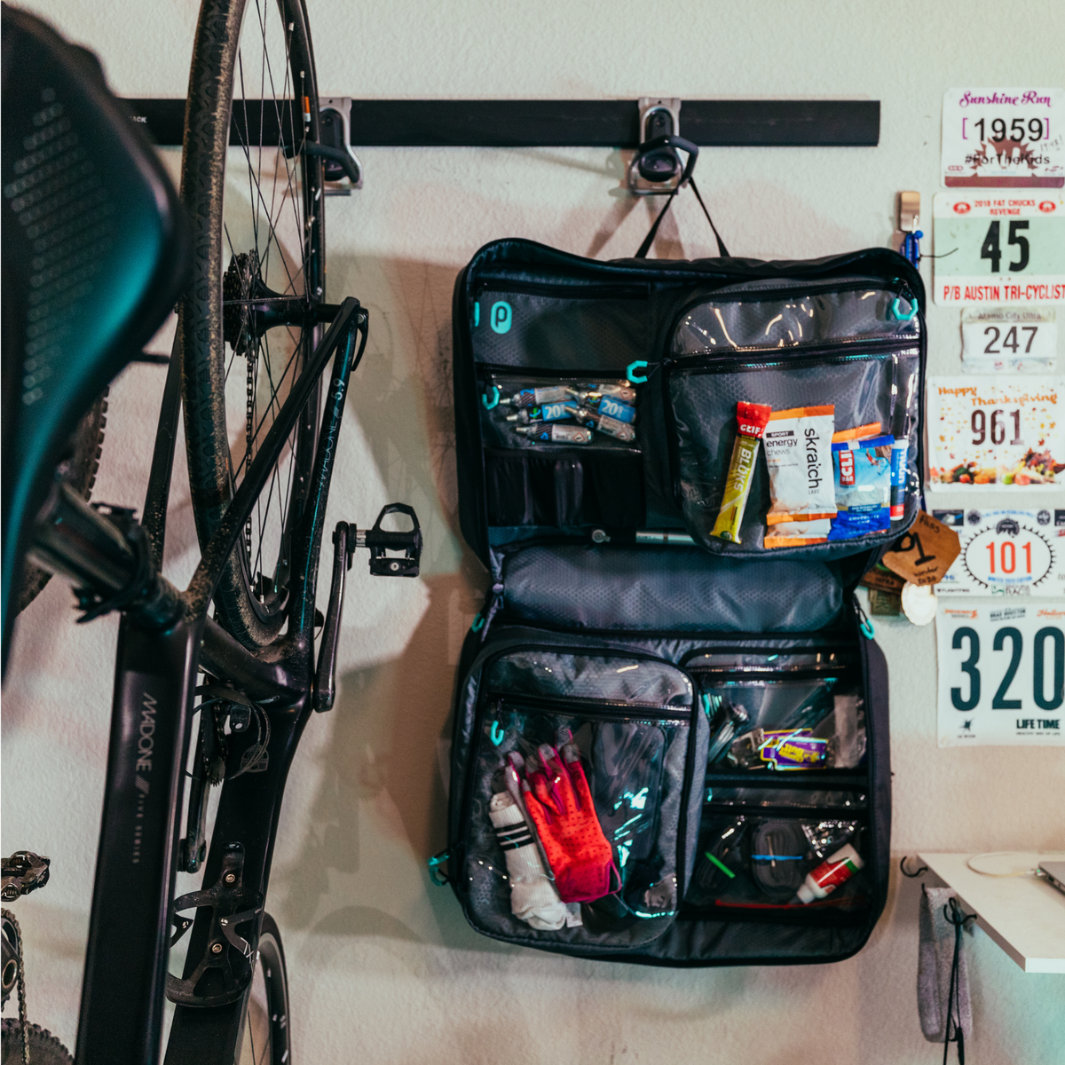 Parc cycling gear bag hanging open on garage wall next to road bike. Bike gear bag filled with cycling kit, tools, cycling nutrition products, mtb gear and other bike gear. Some pockets are clear plastic and some are mesh. Grey interior bag material.